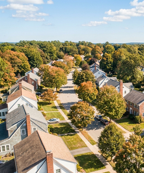 Aerial view of a quiet residential neighborhood with tree-lined streets, detached houses, parked cars, and trees showing autumn colors.